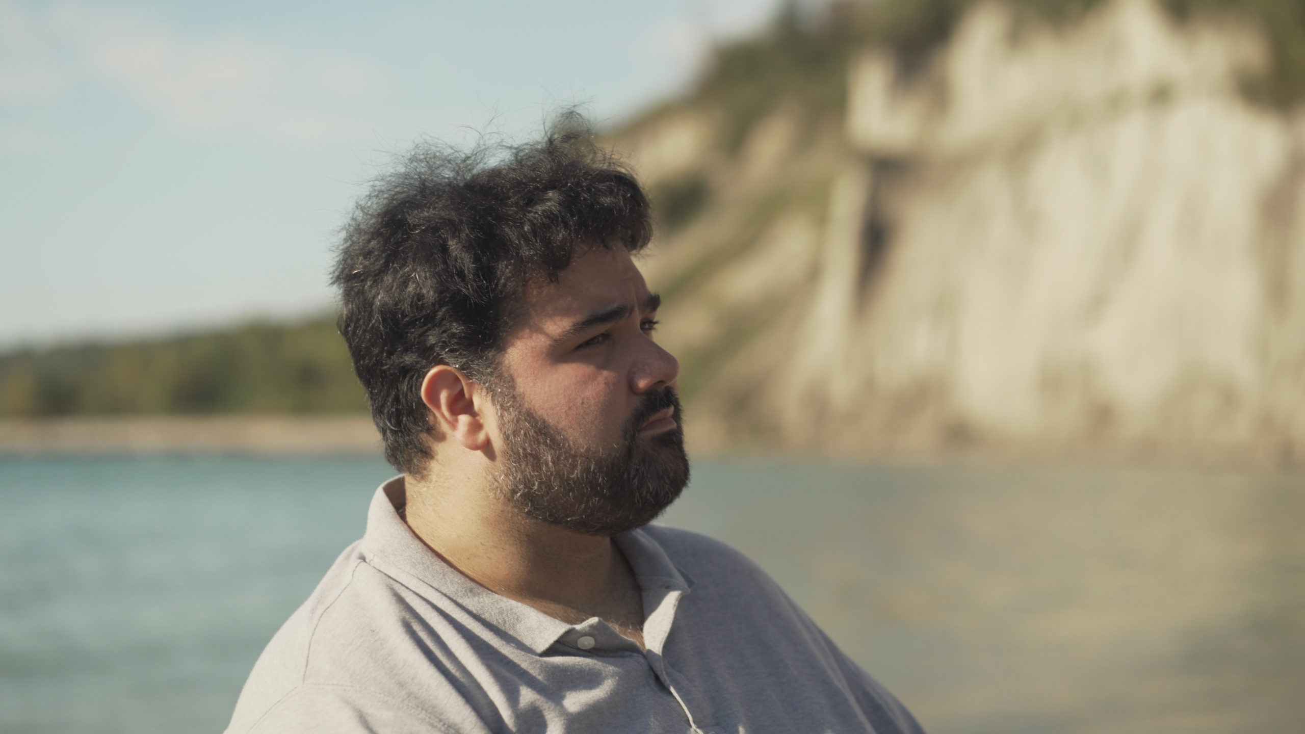 Image of Requiem Soloist Andrew Haji, who has black hair, a beard, and is wearing a white polo, staring off to the right with the Scarborough Bluffs and Lake Ontario in the background.