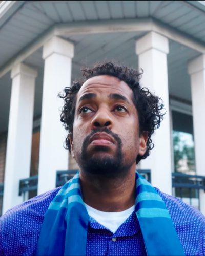 A closeup photo of baritone Justin Welsh. He is a black man with short hair. He is wearing a purple shirt and blue scarf. He is looking out of frame and appears to be distressed. There is a house in the background.