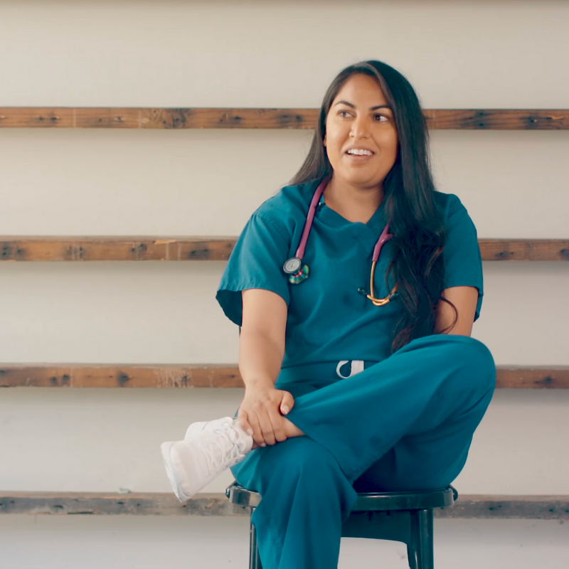 Photo of documentary subject Nadia Vasdani, seated and smiling. She has brown hair and is wear medical scrubs and a pink stethoscope around her neck.