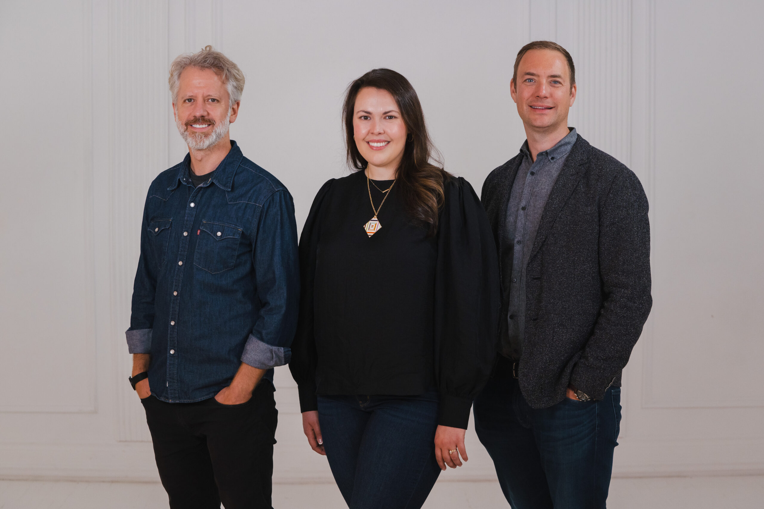 Photo of Founding Artistic Director, Joel Ivany, General Director Robin Whiffen, and Chair of the Board of Directors March Chalifoux. They are all standing together smiling in front of a white wall. Joel is a white man with grey heard and beard. He is wearing a denim shirt and black pants. Robin is a White woman with shoulder-length brown hair. She is wearing all black and a pendant necklace. Marc Chalifoux is a white man with short blond hair. He is wearing a blue shirt underneath a black blazer and with black pants.